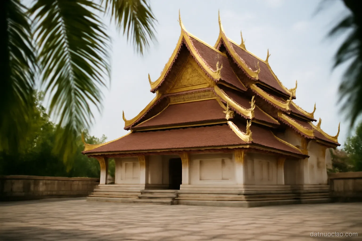 A tiered temple roof with gilded edges seen from a stone courtyard, palm fronds at the edge of frame