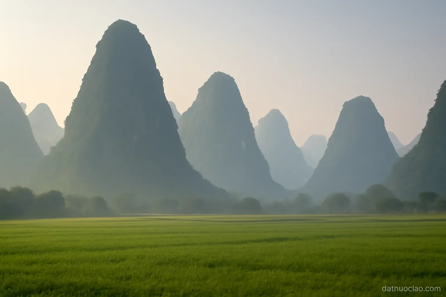 Steep karst limestone peaks rising behind a flat green field, morning haze softening the background